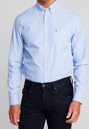 Mężczyzna ubrany w jasnoniebieską koszulę na guziki z kieszenią na piersi oraz ciemnoniebieskie dżinsy, stojący na tle jednolitego szarego tła.