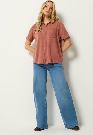 Femme blonde debout contre un mur blanc, portant une chemise rose fanée à boutons, un jean large bleu et des ballerines marron foncé, les mains dans les poches.