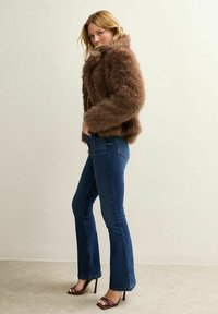 Veste en fausse fourrure marron avec un large col, cintrée à la taille. Associée à un jean évasé bleu foncé et des sandales à talons. Texture lisse.