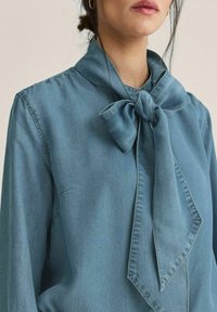 Lyseblå denimbluse med en stor sløjfe ved kraven, der har en skræddersyet design og blød tekstur, som komplementerer den afslappede stil.