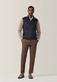 Navy Weste über einem hellbraunen Pullover, braune Hose mit umgeschlagenen Säumen und beigen Schuhen, vor einem neutralen Hintergrund stehend.