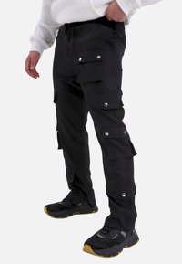 Schwarze Cargo-Hosen mit mehreren Seitentaschen, Druckknöpfen, elastischem Bund und schmaler Passform. Kombiniert mit schwarzen und gelben Sportschuhen.