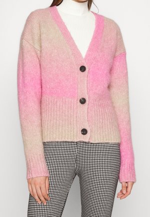 Zachte gebreide cardigan met roze en beige kleurvlakken, drie zwarte knopen, gedragen over een witte coltrui en gecombineerd met zwart-witte pied-de-poule broek.