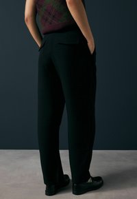 Juoda pritaikyta kelnių su tiesiu kirpimu, minkšta tekstūra ir šoninėmis kišenėmis, derinamos su juodais loafers. Matomas nugarinės kišenės detalė.