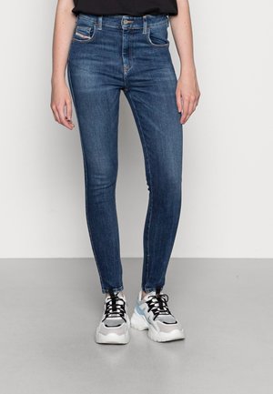 Person iført tætsiddende blå cowboybukser og hvide sneakers med sorte snørebånd stående mod en ensfarvet lys baggrund.