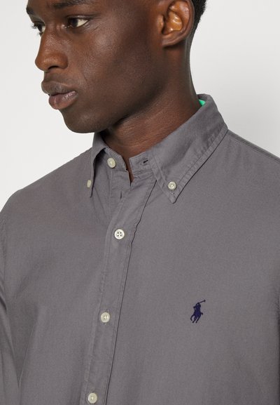 Camisa de botón gris con cuello, que presenta botones blancos y un pequeño logo en azul marino en el pecho. Textura suave, diseño casual.