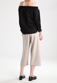 Persona con una blusa nera senza spalle, pantaloni beige a gamba larga e cropped, e mocassini neri, in piedi contro uno sfondo bianco semplice.