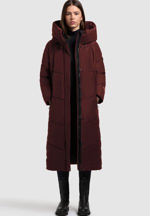 Bordeauxfarbener Puffer-Mantel mit Kapuze, ausgestattet mit einem gesteppten Design, Frontreißverschluss und Druckknopfverschlüssen. Getragen über einem schwarzen Rollkragenpullover und kombiniert mit schwarzen Stiefeln.