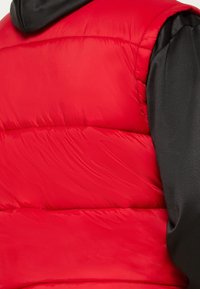 Czerwona pikowana kamizelka z czarnym kapturem, pikowanym wzorem i błyszczącą, gładką fakturą. Widoczne przeszycia i wypełnienie nadają jej miękki, wyściełany wygląd.