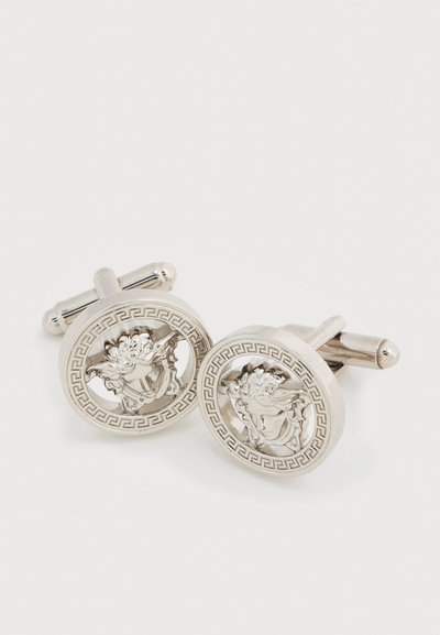 Ασημένια μανικετόκουμπα με κυκλικά σχέδια και πολύπλοκες ανάγλυφες παραστάσεις μιας κλασικής φιγούρας, διακοσμημένα με σχέδιο ελληνικού κλειδιού γύρω από την άκρη.