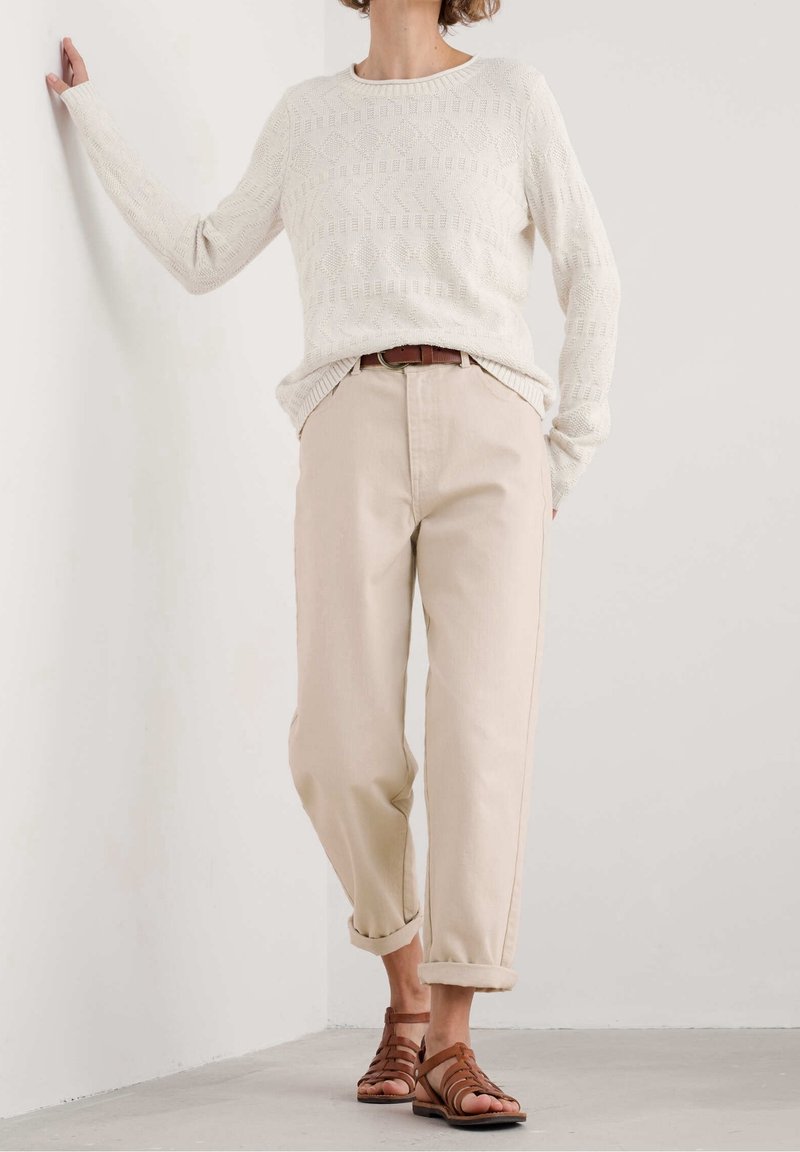 Femme portant un pull texturé blanc à manches longues, un pantalon beige retroussé avec une ceinture marron, et des sandales en cuir marron, debout à l'intérieur.