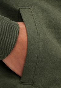 Olíva zöld pulóveranyag texturált felülettel, bordázott mandzsettákkal és oldalzseb részlettel, amely kiemeli a dizájnt.