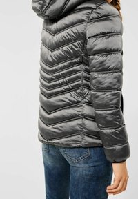 Grå puffjacka med chevronmönster, huva och dragkedjedetaljer; tillverkad av glänsande, vadderat material; kombinerad med blåjeans.