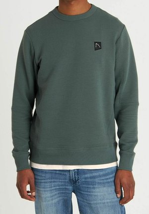 Mand iført en ribbet grøn sweatshirt med et lille sort logomærke på brystet, blå jeans og en synlig hvid skjorte underneden.