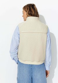 Gilet en polaire beige avec une surface texturée, présentant un col de couleur claire, des poches latérales et un ourlet côtelé, porté sur une chemise à rayures bleues.