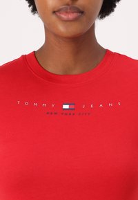 Rød bomulls t-skjorte med rund hals og en logo med et flaggdesign og teksten "TOMMY JEANS NEW YORK CITY" i hvit og blå.