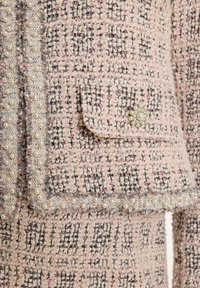 Tweedjas in zachtroze met een zwart-wit frekwentpatroon. Voorzien van parelrand en een decoratieve knop op de linkerzak. Textuurstof.