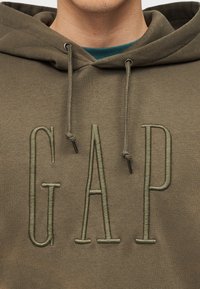 Άτομο με φούτερ σε ελιά πράσινο χρώμα με μεγάλα κεντημένα γράμματα "GAP" στην πρόσοψη και κορδόνια σε ελιά που κρέμονται από τη κουκούλα.