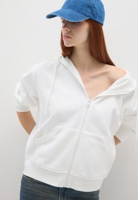 Sudadera blanca con cremallera y corte oversized, que incluye capucha, bolsillos frontales y mangas cortas. Combinada con una gorra de denim azul.
