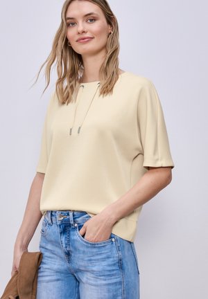 Frau mit lockerem blondem Haar trägt ein hellbeiges, kurzärmeliges Oberteil und blaue Jeans, hält eine braune Jacke in einer Hand vor einfachem Hintergrund.