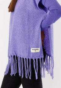 Poncho lavorato a maglia viola con orlo frangiato, etichetta sul lato, vestibilità ampia, texture morbida, indossato sopra pantaloni neri, aperture laterali per maggiore comfort.