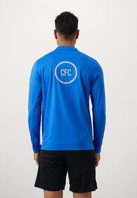 Langærmet bl�å træningsskjorte med et rundt "CFC" logo på ryggen, parret med sorte shorts. Glat stof, tætsiddende design.