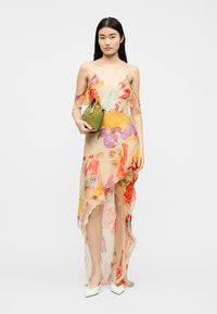 Femme portant une robe florale transparente avec des volants, tenant un petit sac à main vert, et posant devant un fond clair uni.