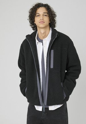Veste en polaire noire avec fermeture éclair, deux poches latérales et une poche sur la poitrine. Présente des accents gris et une finition texturée.