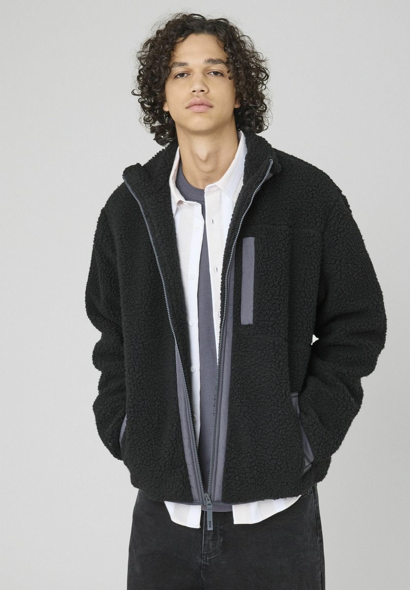 Jeune homme aux cheveux bouclés portant une veste polaire noire sur une chemise blanche et un t-shirt gris, debout les mains dans les poches, fond neutre.