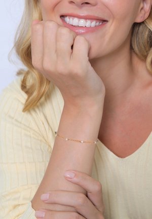 Goldene Kettenarmband, verziert mit kleinen klaren Perlen, getragen am Handgelenk einer Frau. Das Armband ergänzt ein hellgelbes, strukturiertes Oberteil.