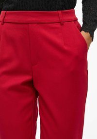 Pantalons ajustés rouges en tissu lisse avec une taille structurée, des poches sur les côtés et des détails de pinces sur le devant.