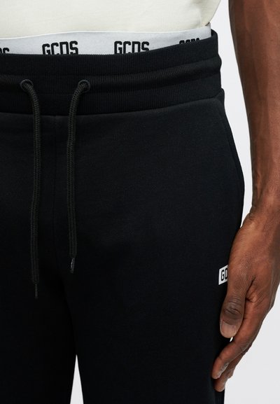 Black sweatpants with elastic waistband and drawstring. Features textured fabric and a small white logo patch. Close-up of hand on waistband.