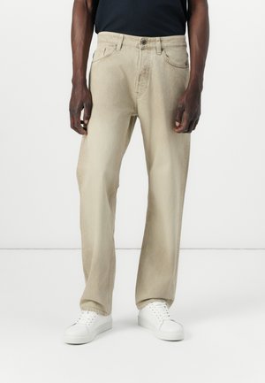 Beige rechte jeans met vijf zakken, subtiele textuur en een knoopsluiting, gecombineerd met witte sneakers en een zwart shirt.