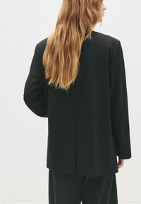 Personne avec de longs cheveux blonds portant un blazer noir et un pantalon noir, de profil contre un fond blanc uni.