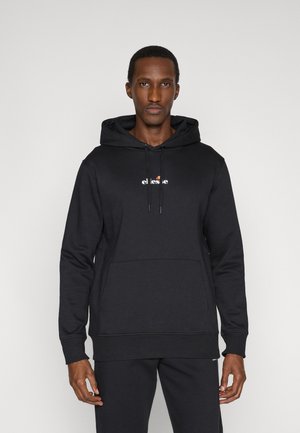 Mann trägt schwarzen ellesse Hoodie mit Fronttasche und Kordeln, steht vor hellem einfarbigem Hintergrund.