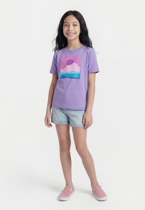 Uśmiechnięta dziewczyna z długimi ciemnymi włosami, ubrana w fioletowy t-shirt z nadrukiem, jasne dżinsowe szorty i różowe buty wsuwane, stojąca na białym tle.