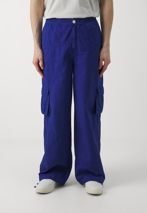 Pantalons cargo bleus à coupe décontractée, avec poches latérales, une taille élastique et une texture de tissu lisse. Associés à des baskets blanches.
