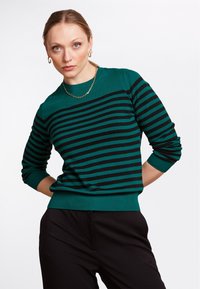 Pull teal avec des rayures horizontales noires, col rond et poignets côtelés. Associé à un pantalon noir à texture lisse et à coupe décontractée.