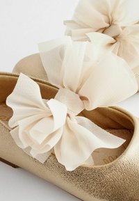 Guldiga platta skor med strukturerat material, med stora beige tulle-bågar framtill. Sula är slät och rundad.