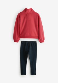 Felpa rossa con colletto alto e una toppa quadrata sul retro, abbinata a leggings blu scuro. Entrambi i capi sono realizzati in tessuto morbido.