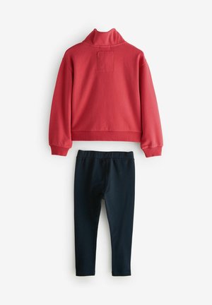 Röd sweatshirt med hög krage och en fyrkantig patch på baksidan, matchad med mörkblå leggings. Båda plaggen är tillverkade av mjukt tyg.