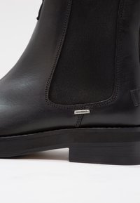Bottine Chelsea en cuir noir avec panneau latéral élastique, talon bas et petite étiquette métallique indiquant "SHABBIES" près de la semelle.