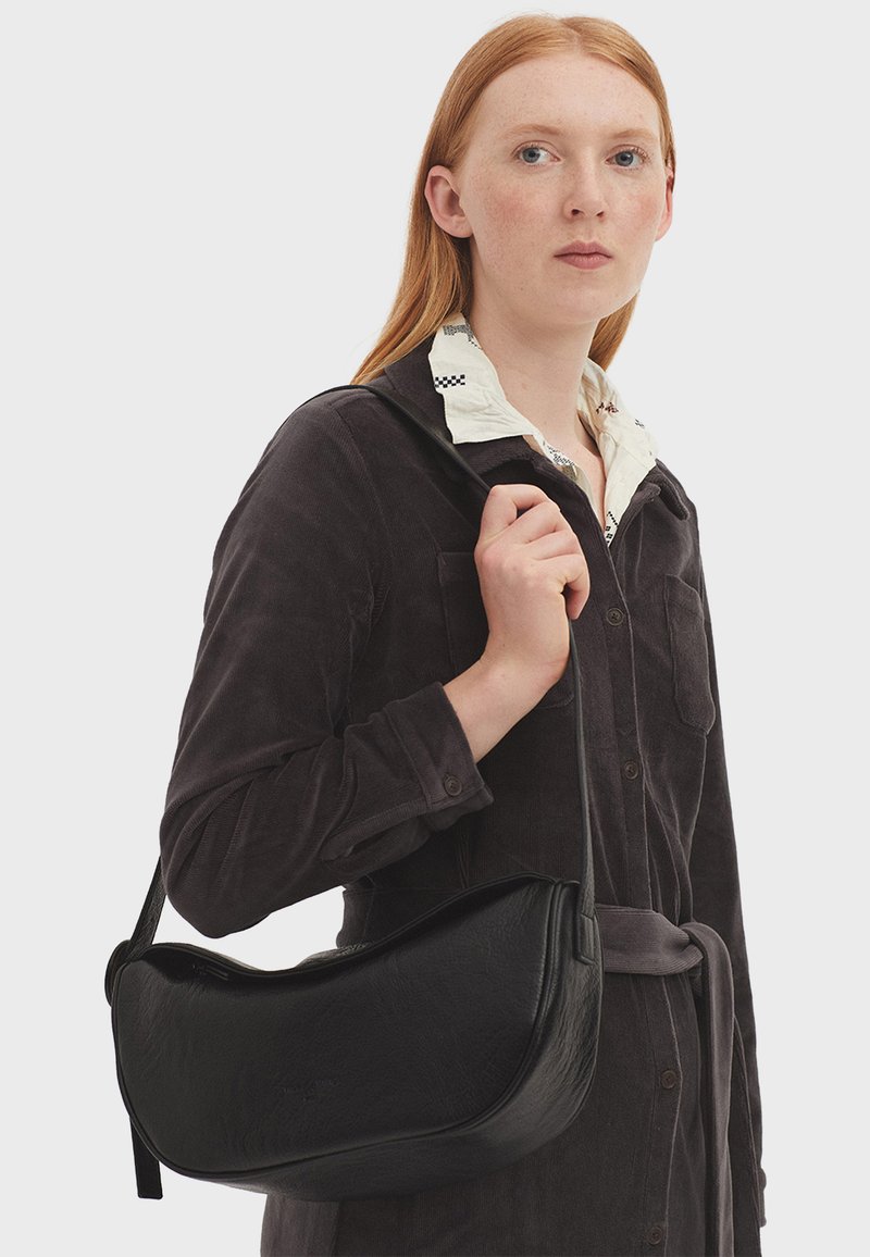 Borsa a tracolla nera in pelle con una forma curva e una texture liscia, dotata di una lunga tracolla e di un design minimalista; indossata sopra un outfit grigio.