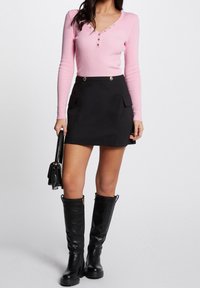 Kobieta ubrana w różową bluzkę z długim rękawem, zapinaną na guziki, czarną mini spódnicę z przednimi kieszeniami, czarne kozaki do kolan, trzymająca czarną torebkę.