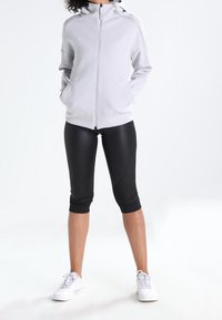 Chaqueta de cremallera gris claro con cuello alto, combinada con leggings capri negros con acentos de malla. Zapatillas deportivas blancas completan el look.