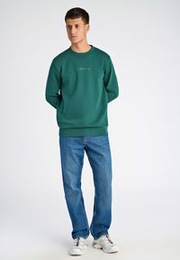 Grön sweatshirt med ribbad textur och rund halsringning, med en logotyp i mitten. Bärs med blå jeans och vita sneakers.
