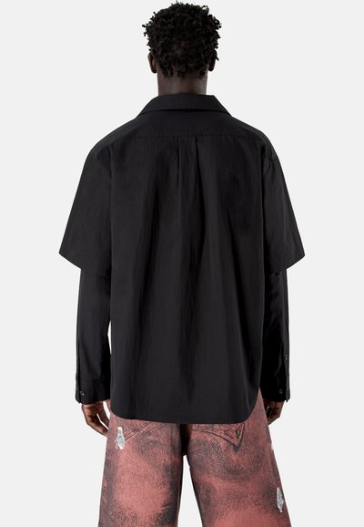 Hombre con cabello corto y rizado que lleva una camisa negra de manga larga y holgada debajo de una camisa negra de manga corta y pantalones cortos rojos con estampado, visto de espaldas.