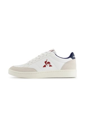 Sneaker basse blanche avec des accents en daim beige, logo de coq rouge sur le côté, doublure de col bleue marine et lacets blancs sur une semelle blanche.