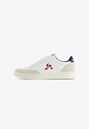 Sneaker bianca bassa con dettagli in suede beige, logo del gallo rosso sul lato, fodera del colletto blu navy e lacci bianchi su una suola bianca.