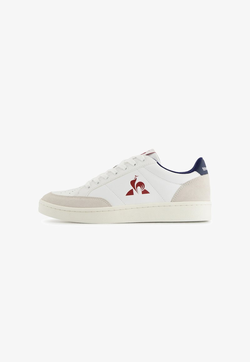 Witte lage sneaker met beige suède accenten, rood hanenlogo op de zijkant, marineblauwe kraagvoering en witte veters op een witte zool.